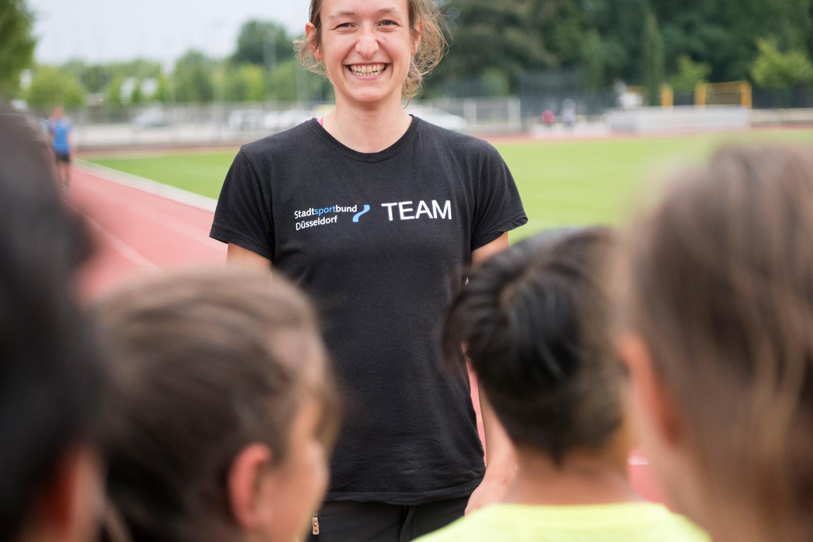 Lächelnde Trainerin auf der Laufbahn, umgeben von Kindern in Sportkleidung, die ihr gespannt zuhören.