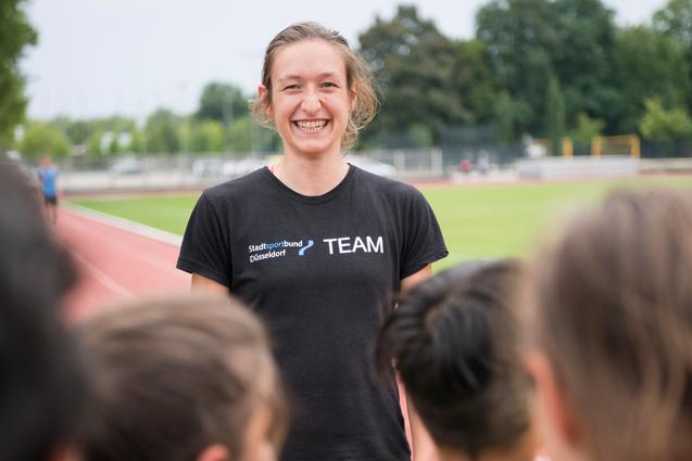 Lächelnde Trainerin auf der Laufbahn, umgeben von Kindern in Sportkleidung, die ihr gespannt zuhören.