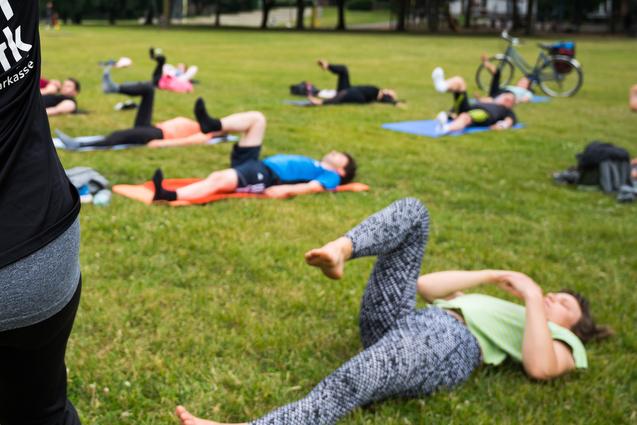 Trainerin mit schwarzem Shirt leitet Sportübungen im Park; Teilnehmer liegen auf Matten und trainieren im Hintergrund.
