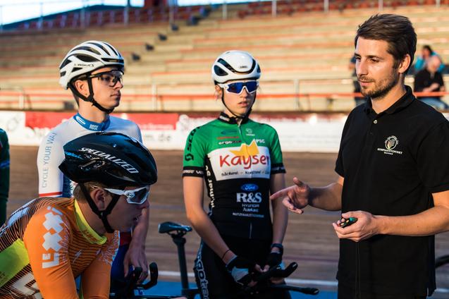 Eine Gruppe junger Radfahrer hört einem Trainer auf einer Radrennbahn aufmerksam zu.