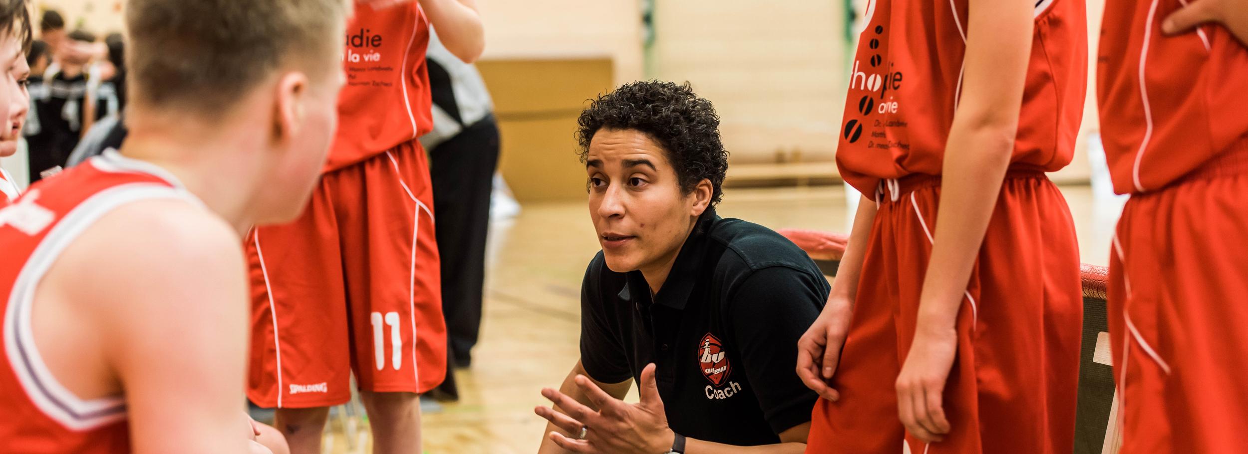 Basketballtrainerin erklärt Taktik während einer Auszeit zu Jugendlichen in roten Trikots auf dem Spielfeld.